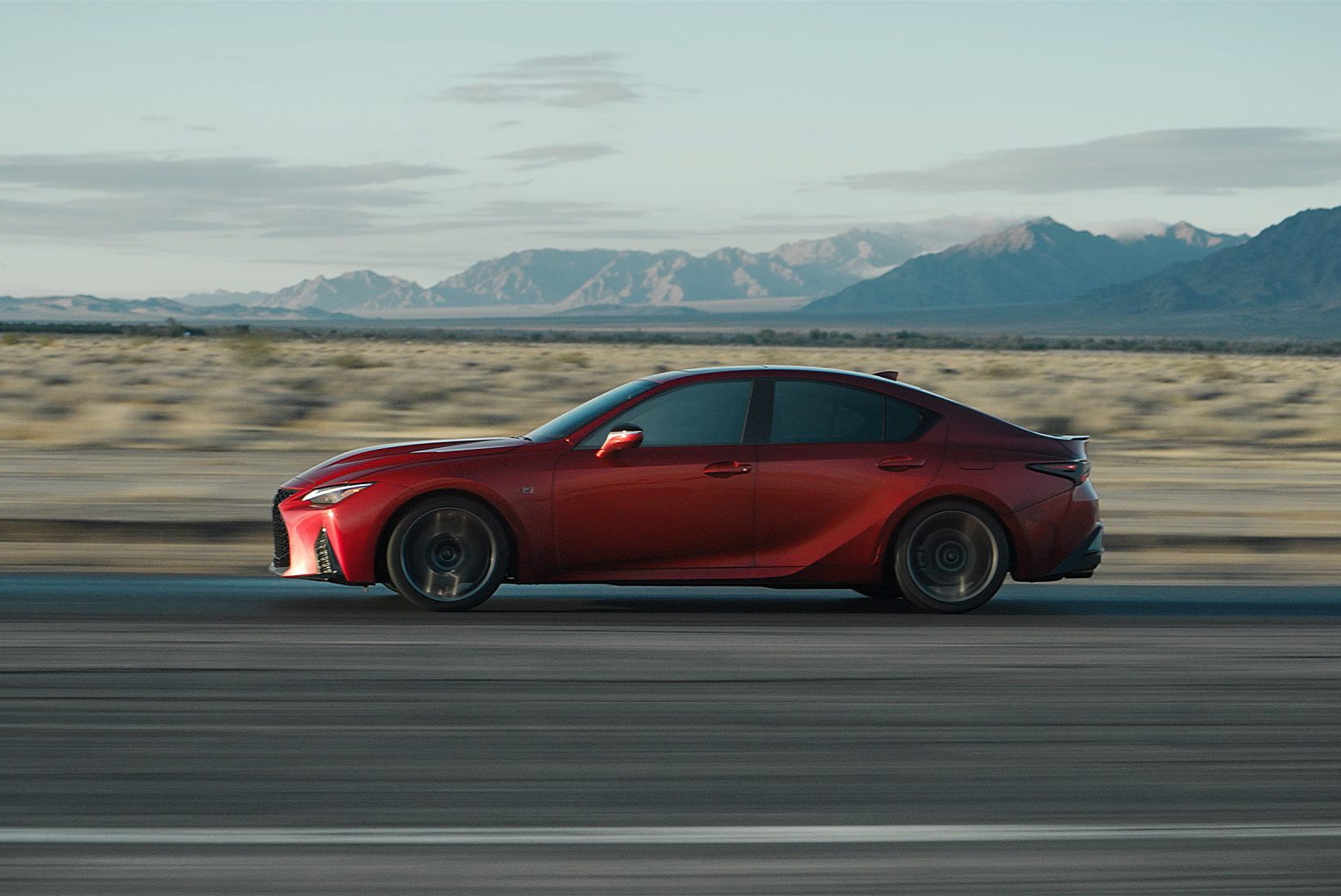 A red sport sedan driving at speed on an open desert highway, shown in side profile with blurred road motion, mountains in the distance, and a clear sky overhead.