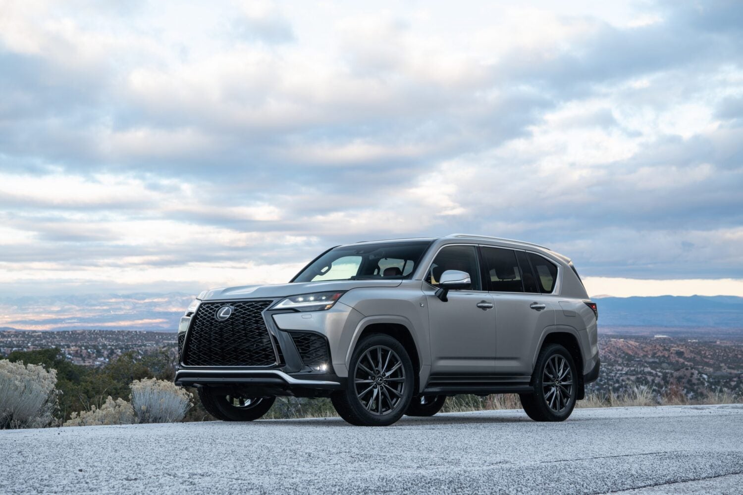 A silver Lexus SUV parked on a paved overlook, shown in three-quarter front view with a large black grille, dark alloy wheels, and a wide landscape and cloudy sky in the background.