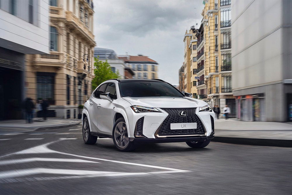 A white Lexus UX compact SUV driving on a city street with European-style buildings in the background.