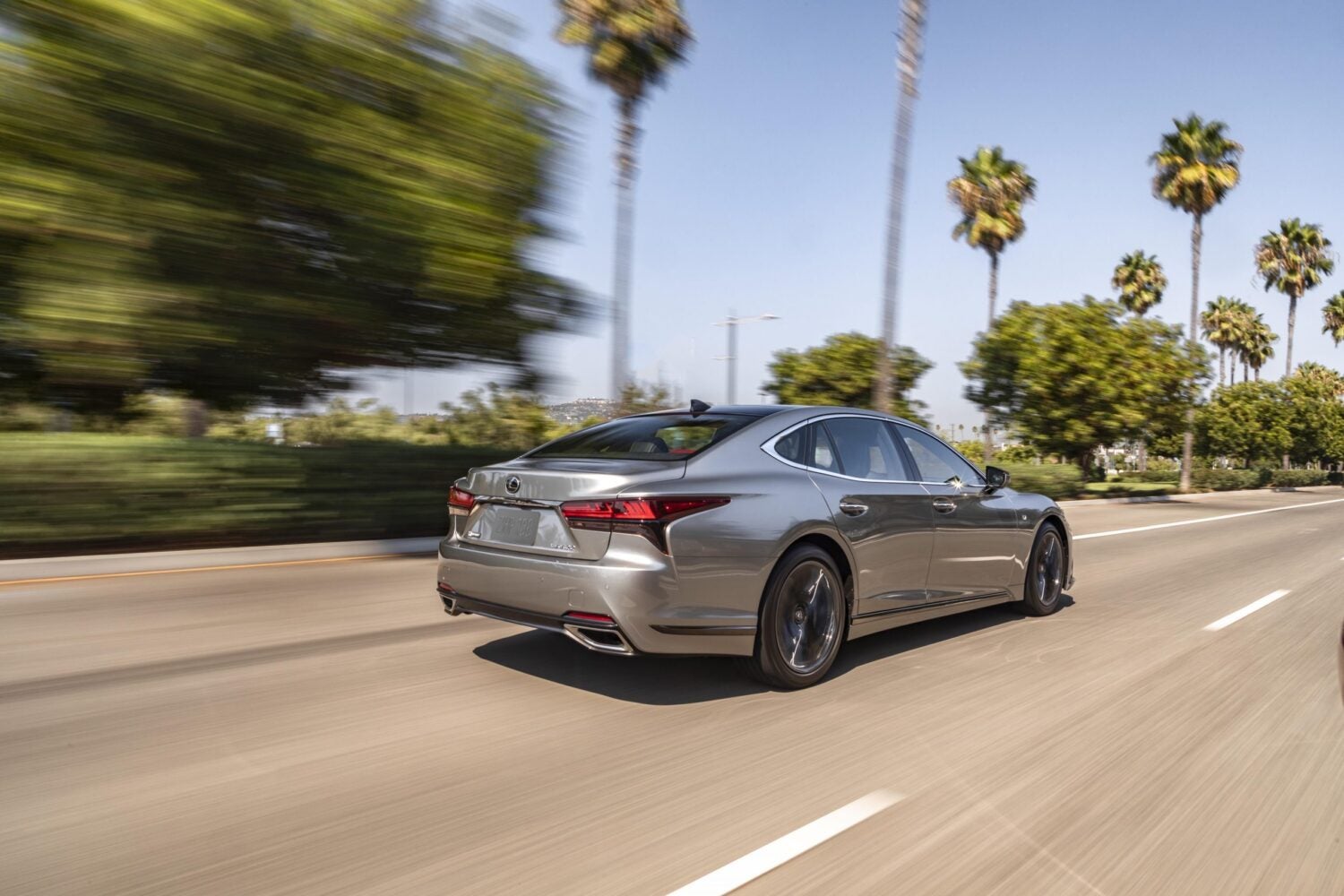 A silver 2026 Lexus LS in Phoenix drives away from the viewer to the right on a highway with palm trees in the background.