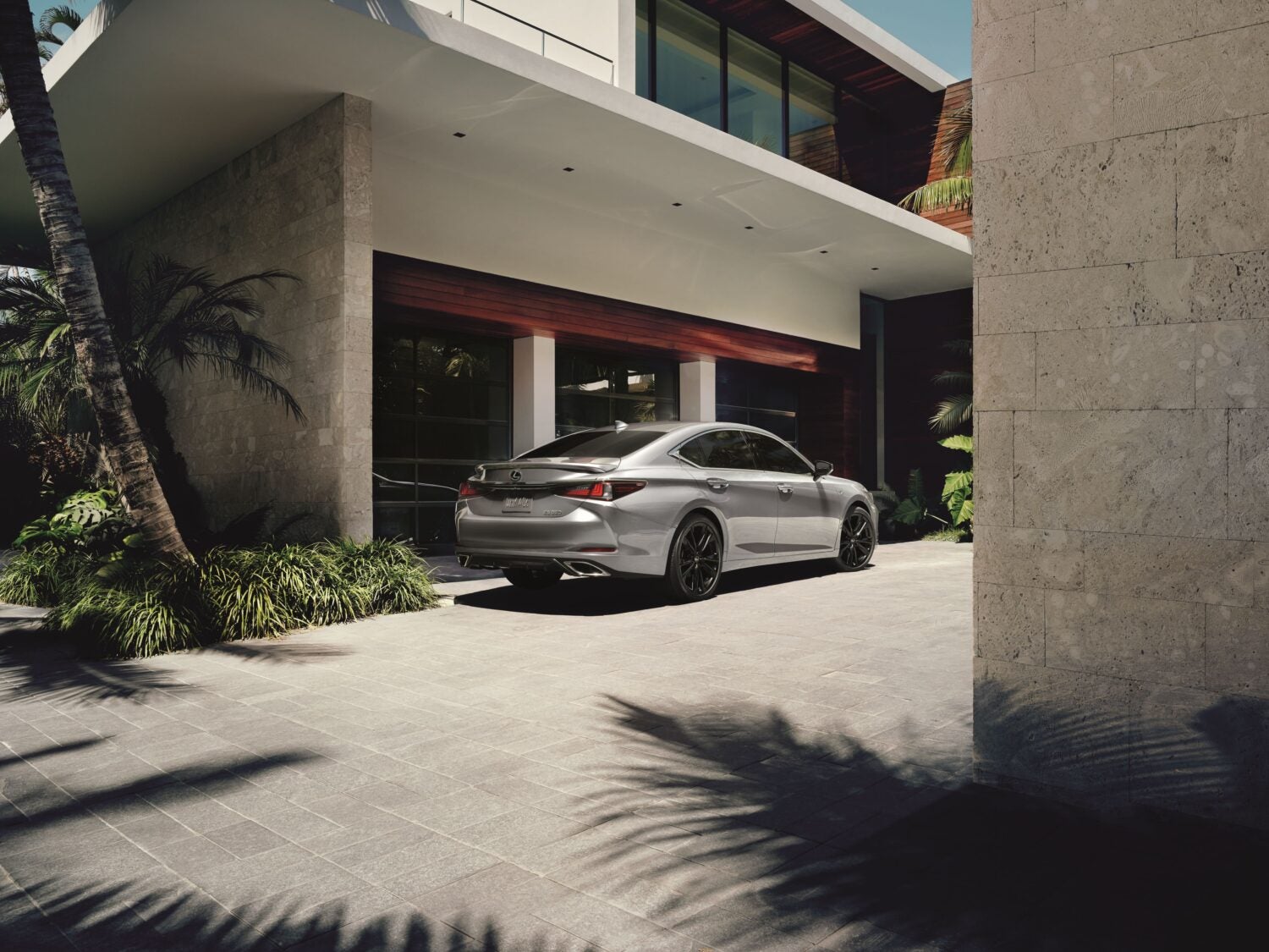 A silver Lexus ES sedan is parked on a paved driveway in front of a modern, multi-story building with stone and wood accents. Palm trees and lush greenery are visible around the building, suggesting a warm climate.