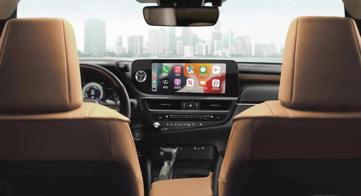 A view from the rear seats of a car, showing the dashboard and the infotainment screen. The screen displays a colorful grid of app icons, and in the background, a cityscape with tall buildings is visible through the front windshield. The car's interior features tan leather seats.
